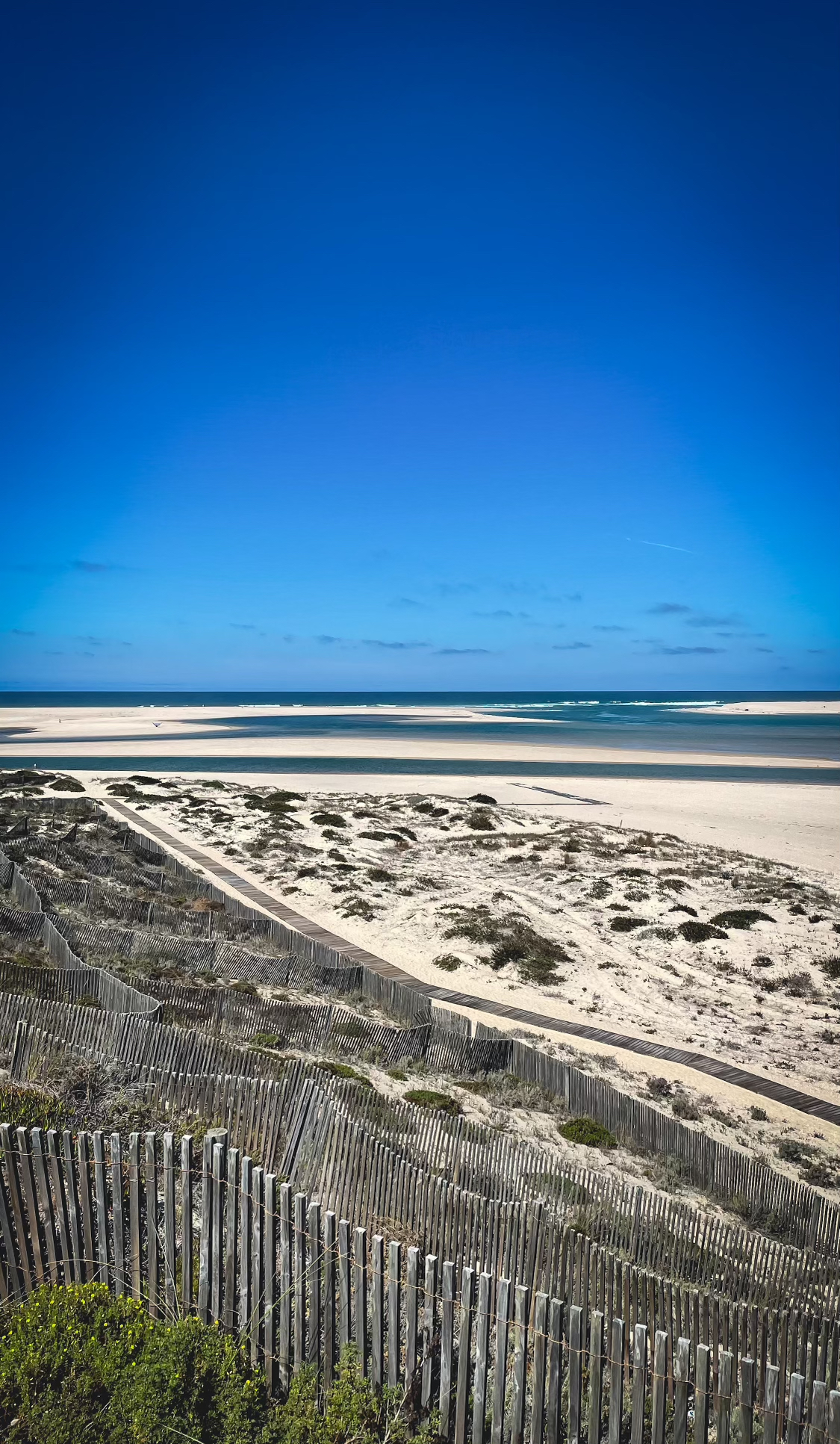 Serene Beach Path: Wooden boardwalk meandering through sand dunes under a clear blue sky, leading to a tranquil beach and turquoise sea, capturing the essence of coastal beauty for a travel blog.