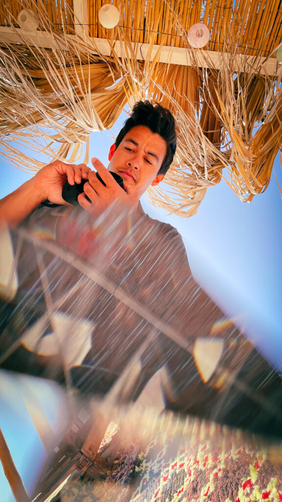 A man under a straw parasol views his phone, with the bright blue sky above and a blurred beach scene below.