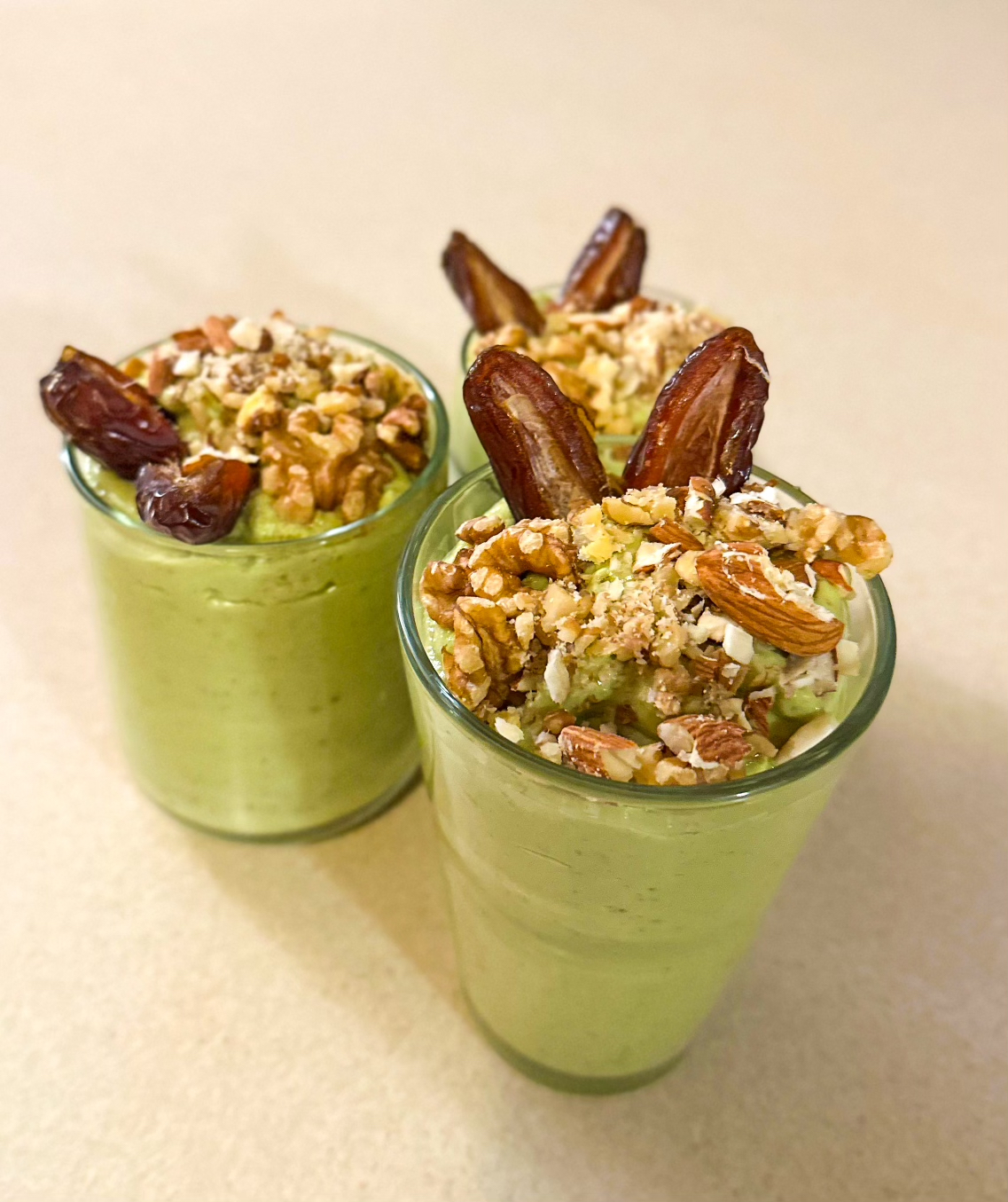 Three glasses of creamy green avocado lassi topped with chopped nuts and dates