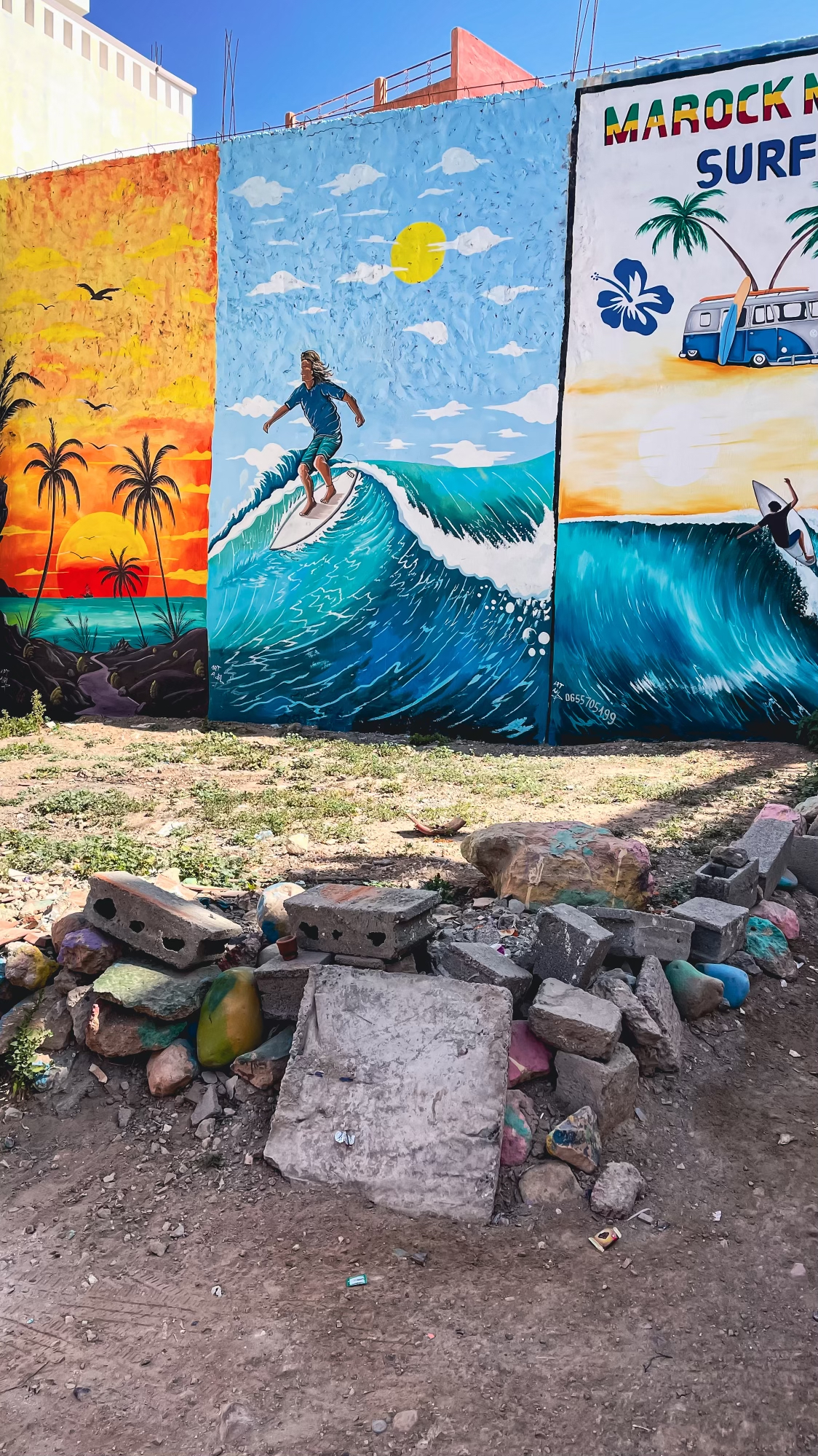 Colorful street art mural in Tamraght, Morocco, depicting a surfer riding a vibrant blue wave with tropical scenery and a retro van, showcasing the local surf culture and artistic spirit.